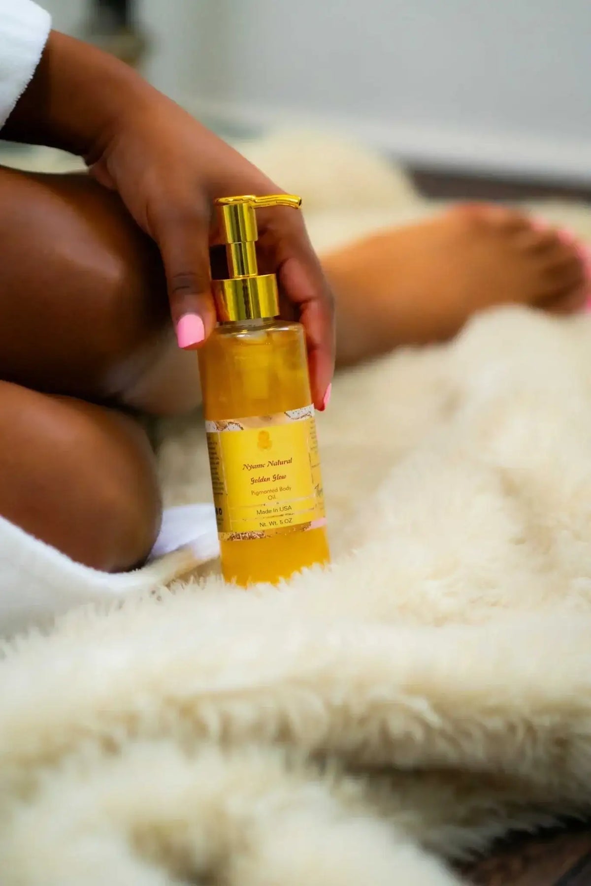 A person holding a bottle of 24k Gold Pigmented body oil, showcasing its luxurious glow and moisturizing properties.