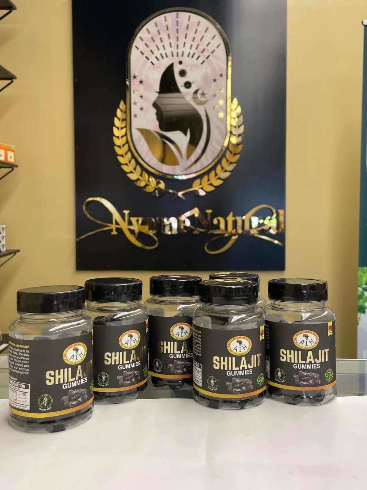 Five bottles of RoyalLuxsLLC Shilajit gummies with black lids on a white surface in front of a decorative sign