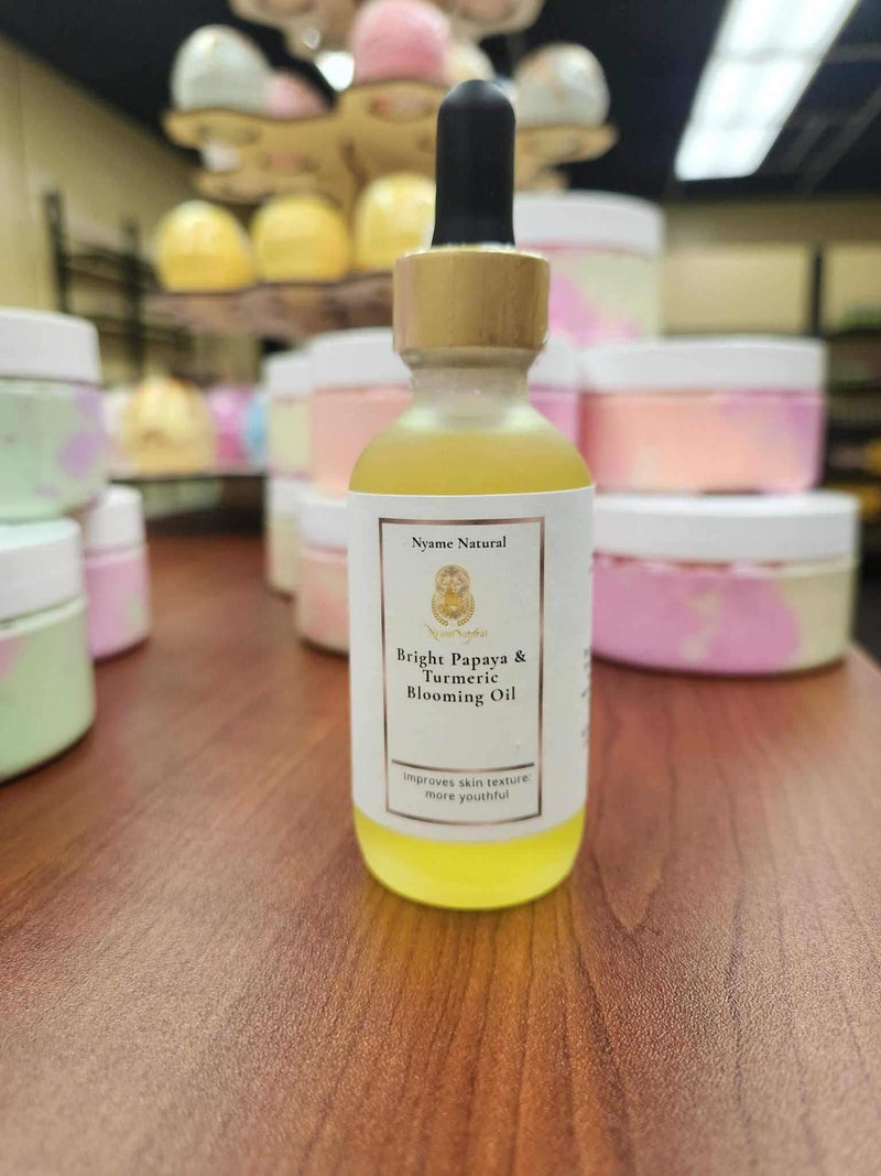 Bright Papaya & Turmeric Blooming Oil in a glass dropper bottle on a wooden surface with colorful bath products in the background.